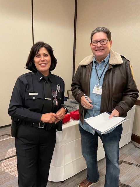 Officer in uniform standing next to a man with glasses holding papers in a conference room setting.